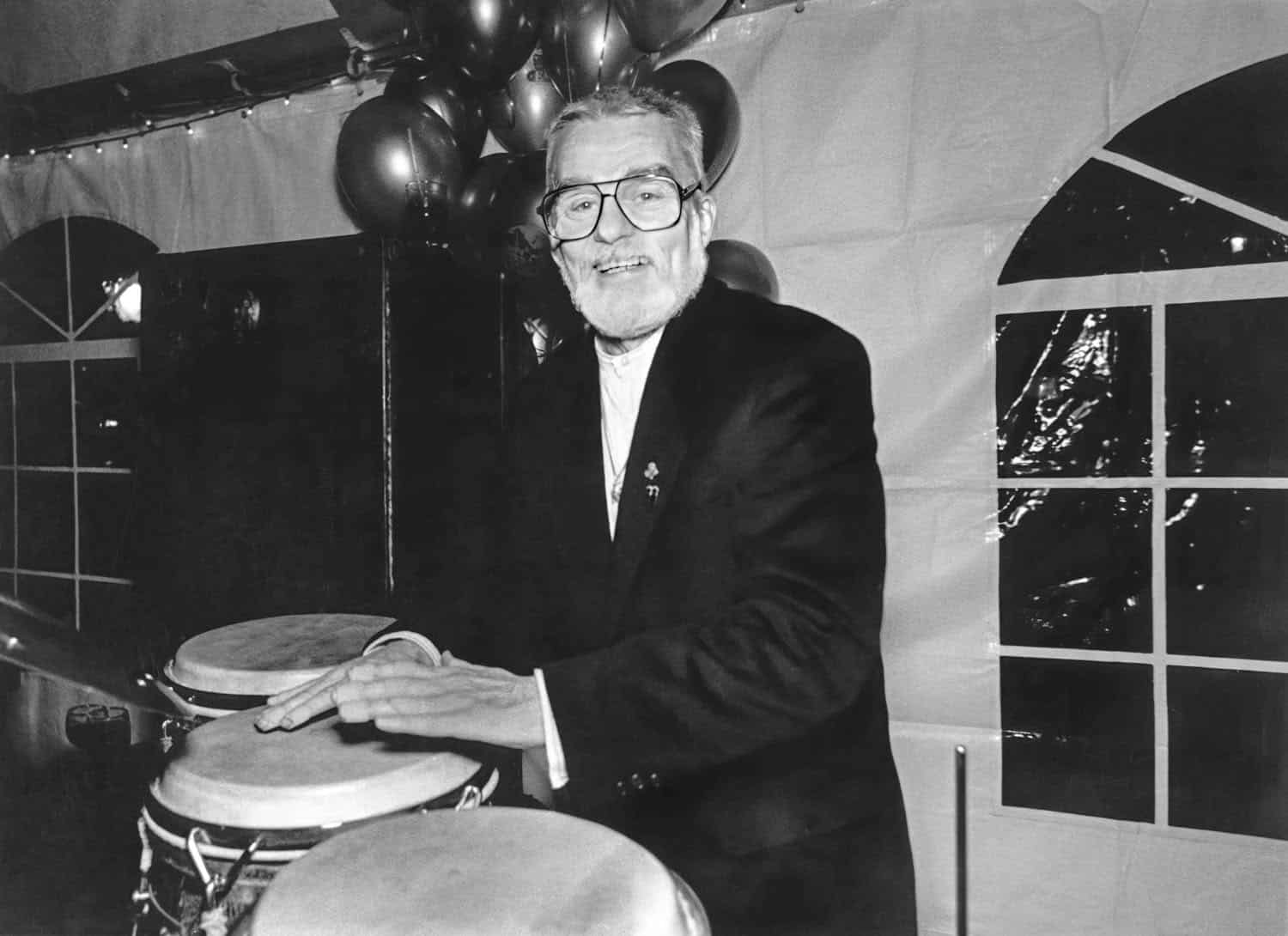 Black-and-white photograph of a matured man with white hair in a large venue tent smiling at the camera. He is wearing a black and white suit and there are balloons behind him.