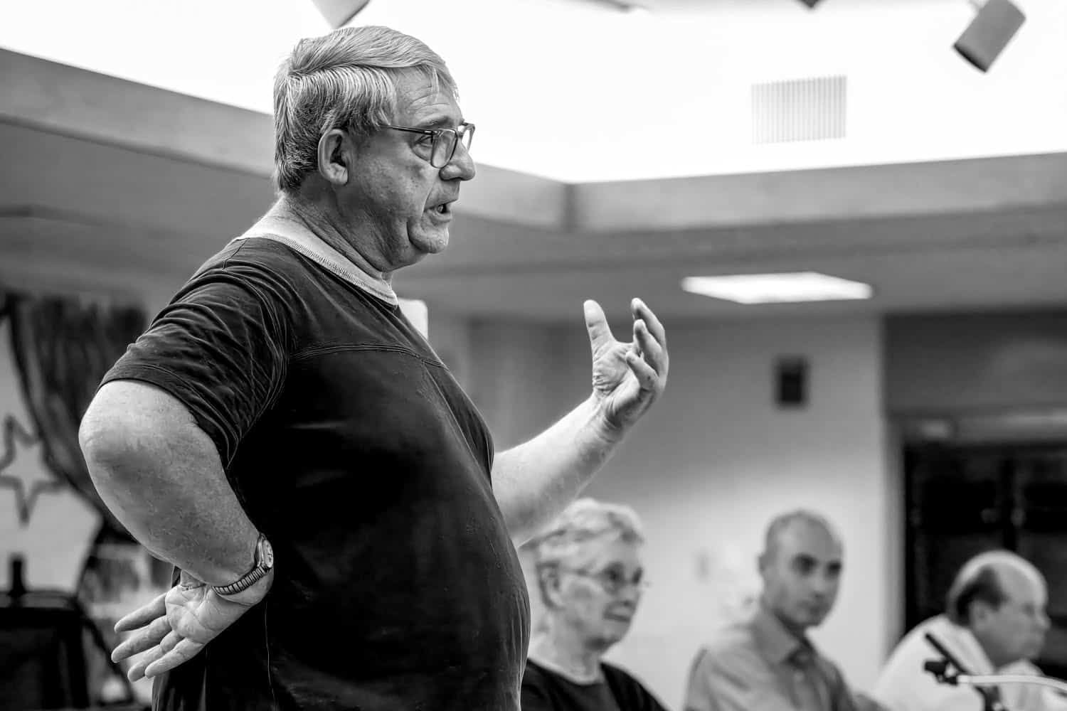 Black-and-white photograph of a man speaking and putting out his arm in an expressive way. He is middle aged or matured, and has graying hair and glasses. He is clean shaven and wearing a black t-shirt. People can be seen in the blurred background.