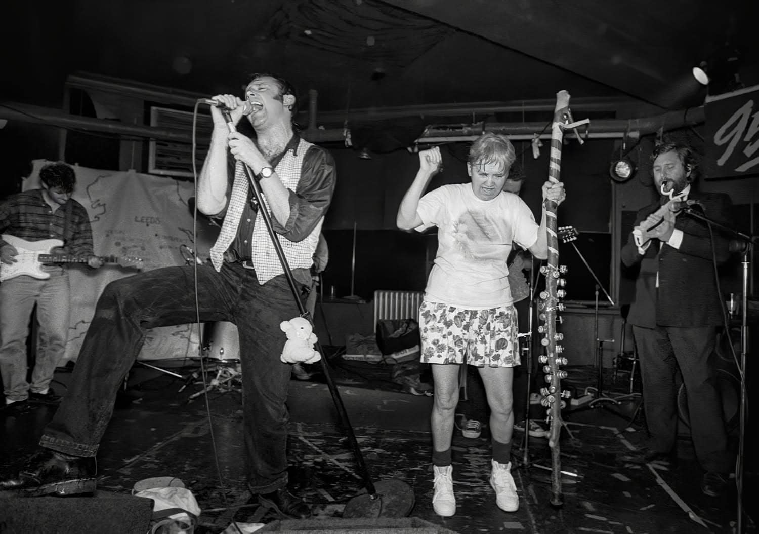 Black-and-white photograph of a band on stage. A man sings into a microphone expressively with his lead propped up. A woman next to him is dancing while holding onto a large stick with bells.