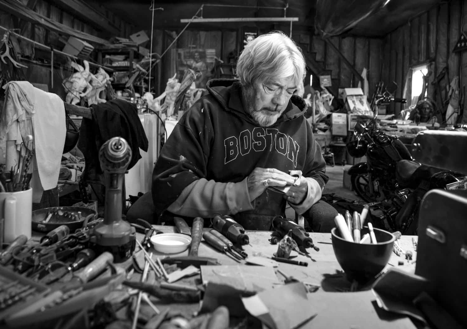 Photograph of a matured man working with tools in a shed or studio. He has white hair and a white beard and is looking down at what he is doing. He has a dark Boston sweatshirt on. 