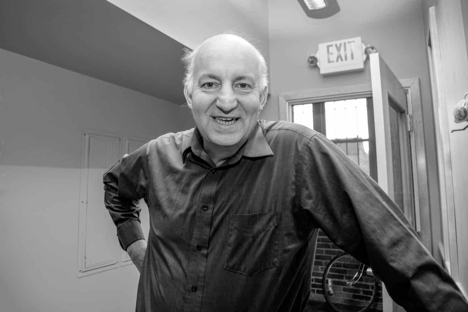 Black-and-white photograph of a middle aged man with white balding hair and a black buttoned up shirt. He is infront of a door with an exit sign. 