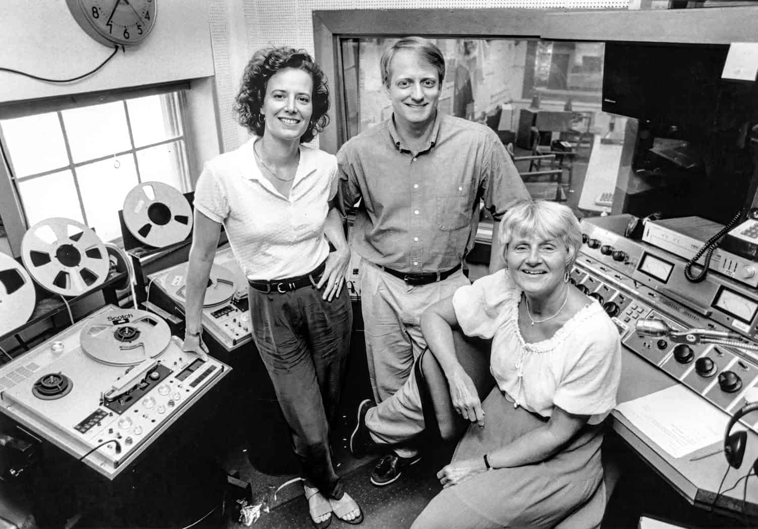 Black-and-white photograph of three people in a radio recording studio. The equipment can be seen around them. On the left is a woman with short curly dark hair wearing a collared shirt and jeans. Next to her is a man with light hair in a buttoned down shirt and pants. Sitting next to them is a matured woman with white or light hair, wearing a white blouse. They all are smiling at the camera.