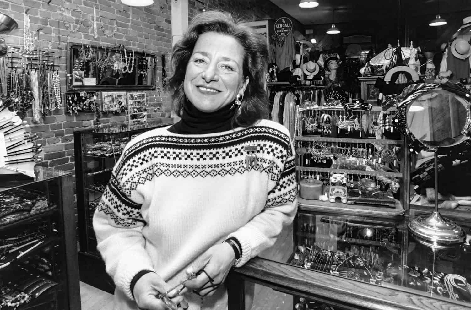 Black-and-white photograph of a woman in the old Country Comforts store at the checkout counter. Her arm is leaning on the counter and she has short wavy brown hair. She is smiling at the camera and wearing a winter sweater.