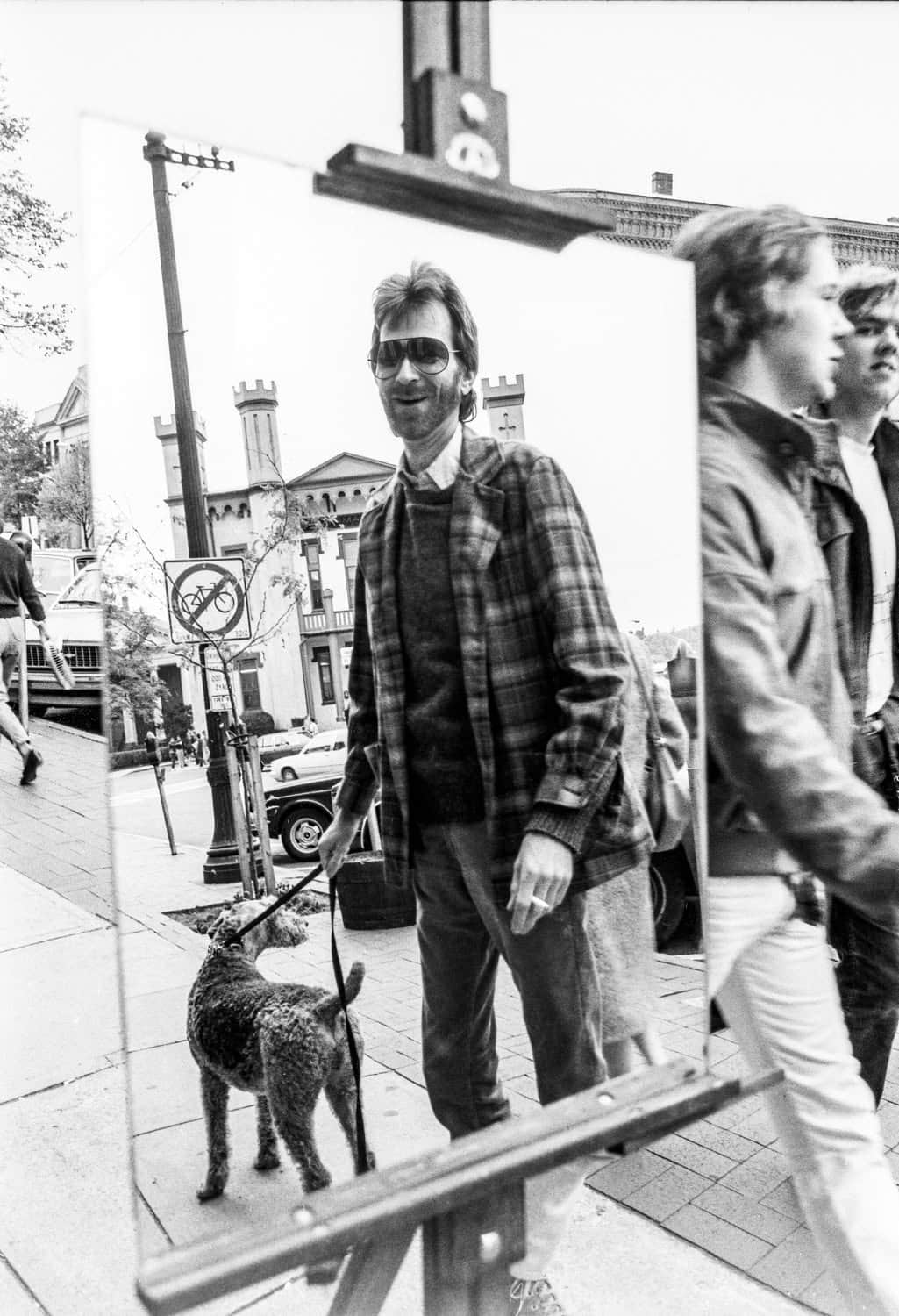 Photograph of a middle aged man walking a dog looking at his reflection in a mirror placed in a canvas stand. The man has short dark hair, sunglasses, is beared, and is wearing a flannel, dress top, and jeans. He is walking on Main Street in Northampton. He smiles at his reflection.