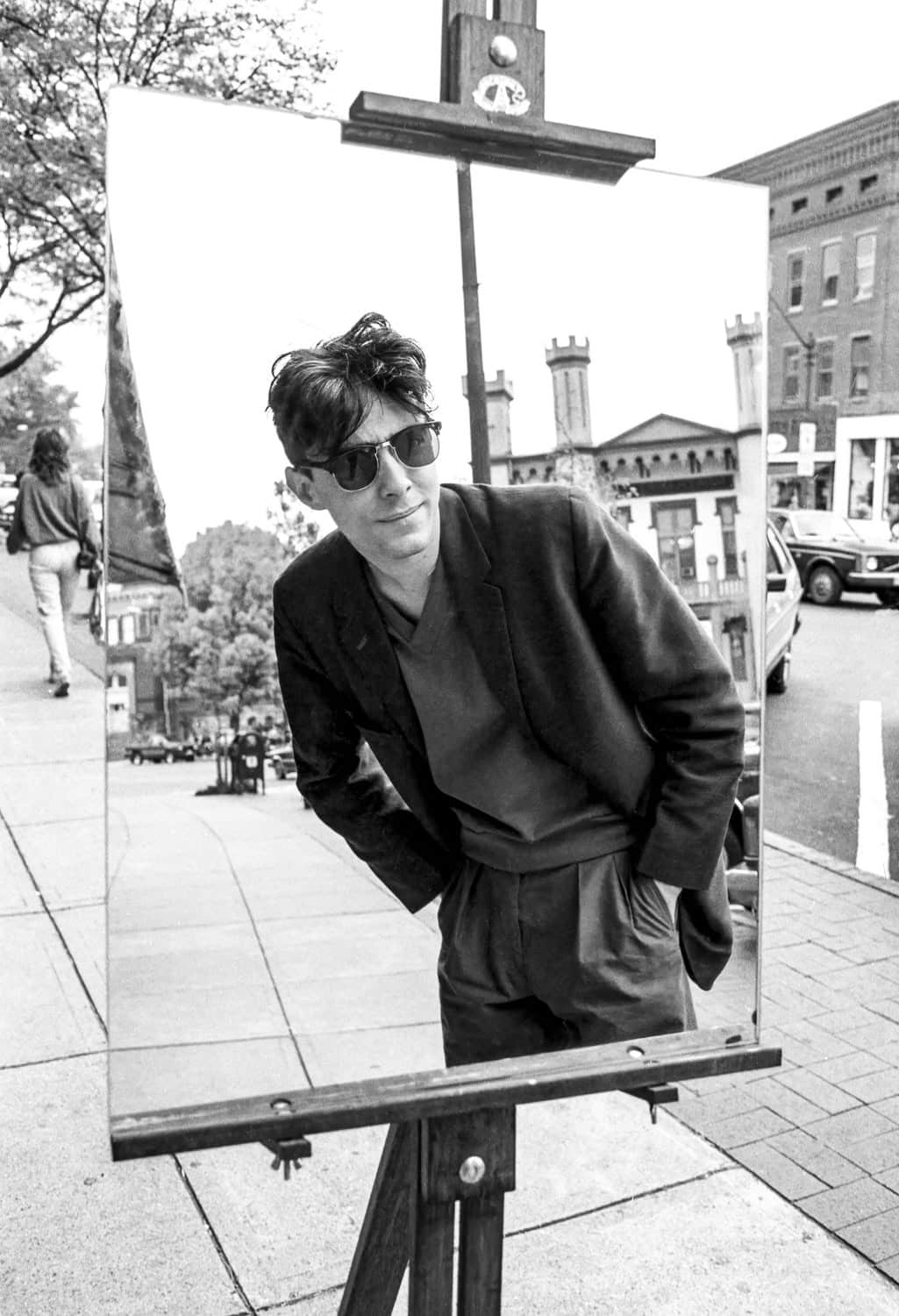 Black-and-white photograph of a young man leaning to see his reflection in a mirror set up on a canvas stand. The man is on Main Street. He has short dark disheveled hair and sunglasses. He is wearing a dark suit jacket and dark dress paints, and is putting his hands in his pants pockets. He is smirking at himself in the mirror.