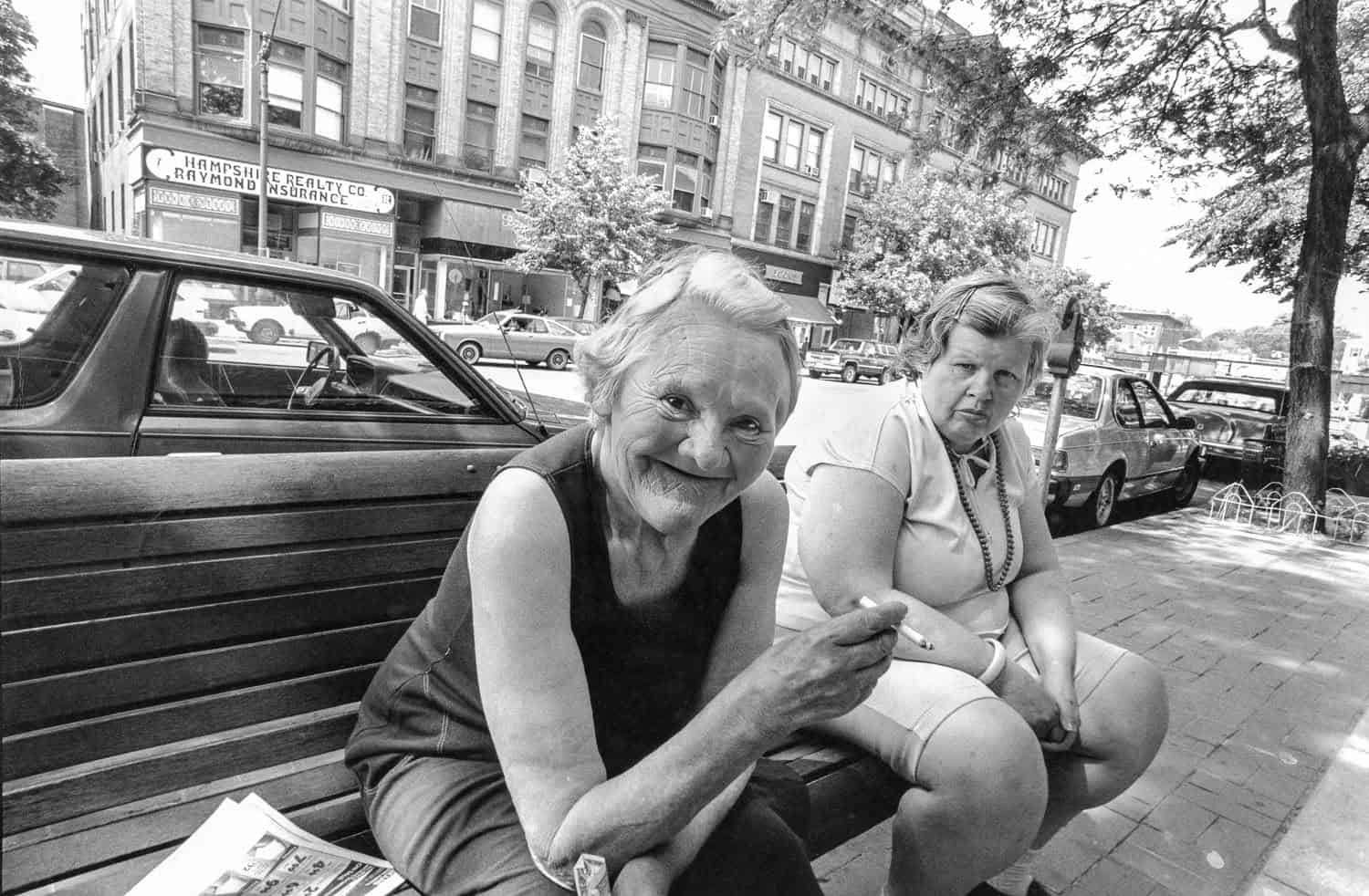 Photograph of two women in downtown Northampton sitting on a bench, storefronts can be seen in the background. The woman closest to the camera is a matured woman in a black tanktop holding a cigarette. Her hair is short and white and she is smiling at the camera. The woman next to her is middle aged and in a light-colored short sleeve top and shorts. Her hair is short and pinned away from her face. She looks neutrally at the camera.
