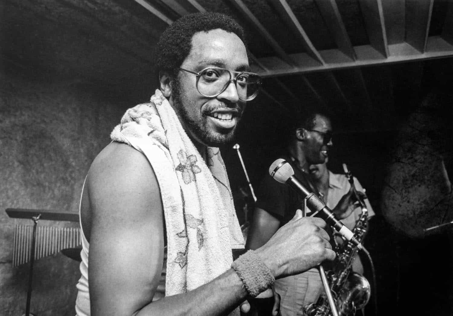 Black-and-white photograph of a man on stage infront of a microphone. He has his face turned toward the camera and is smiling. He is shirtless with a towel draped around his neck. He has a dark beard and short coiled hair. He has large thin framed glasses on. Another man is in the background holding a saxophone facing in another direction.