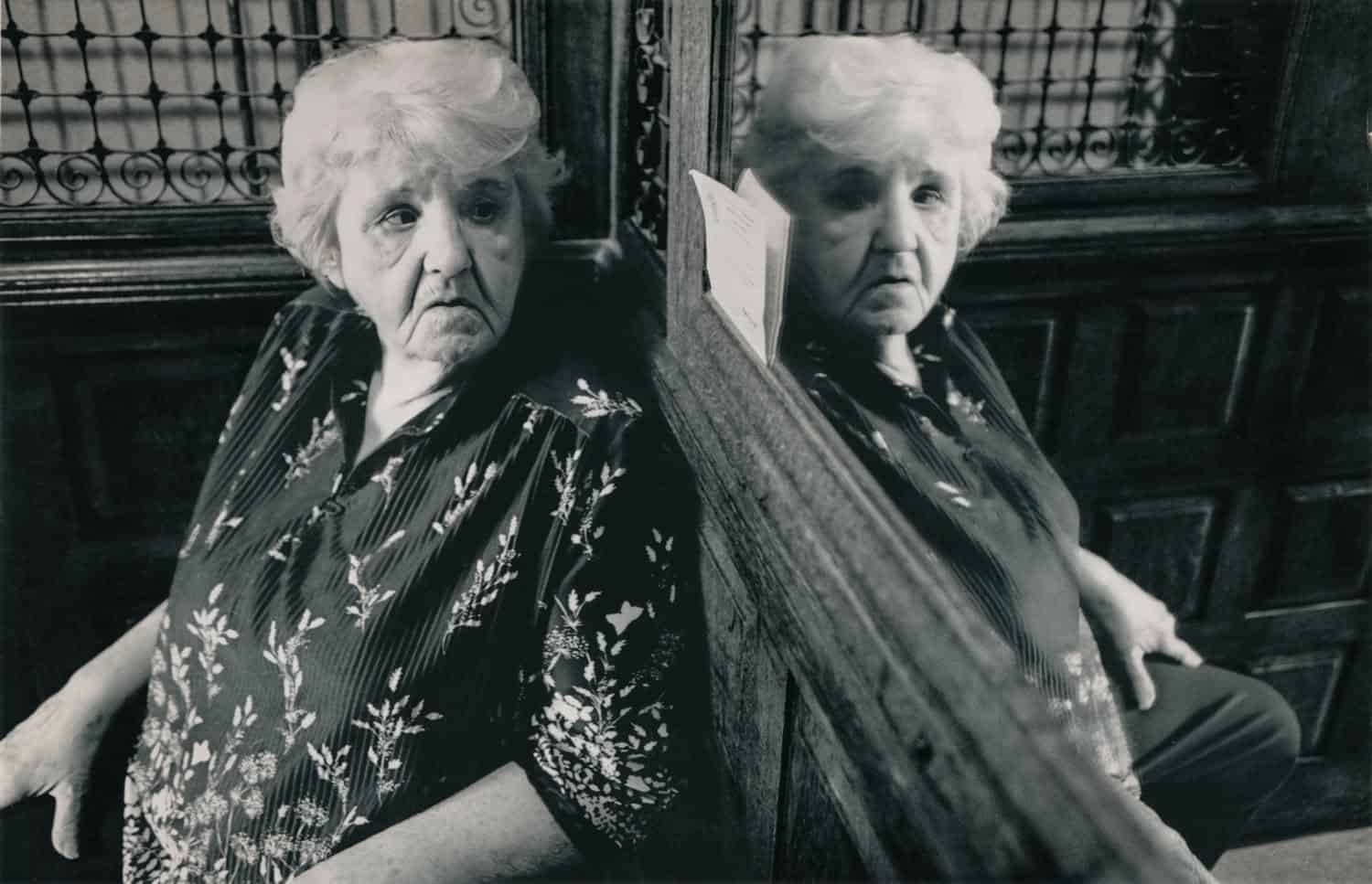Black-and-white photograph of a woman sitting on an old elevator looking at her reflection in a large mirror on the wall. The woman is matured with white short hair and a dark floral top.
