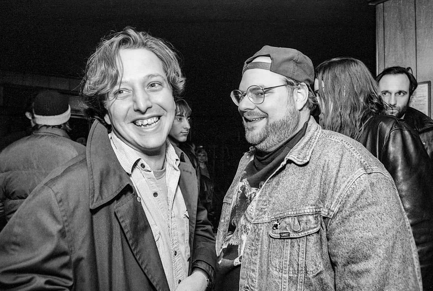 Black-and-white photograph of two young men in their 20s or 30s smiling at the camera. Other people are standing around in the background.