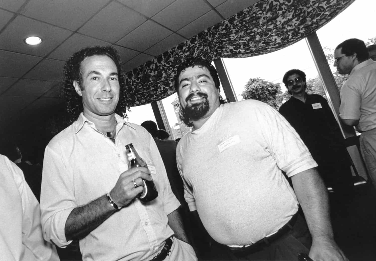Black-and-white photograph of two middle aged men, people are standing around in the background. The man on the left is wearing a white buttoned down shirt and has shoulder length curly hair. He is holding a beer bottle. The man next to him has short hair and a beard, he is wearing a long sleeved t-shirt and has on a name tag. Both men smile at the camera.