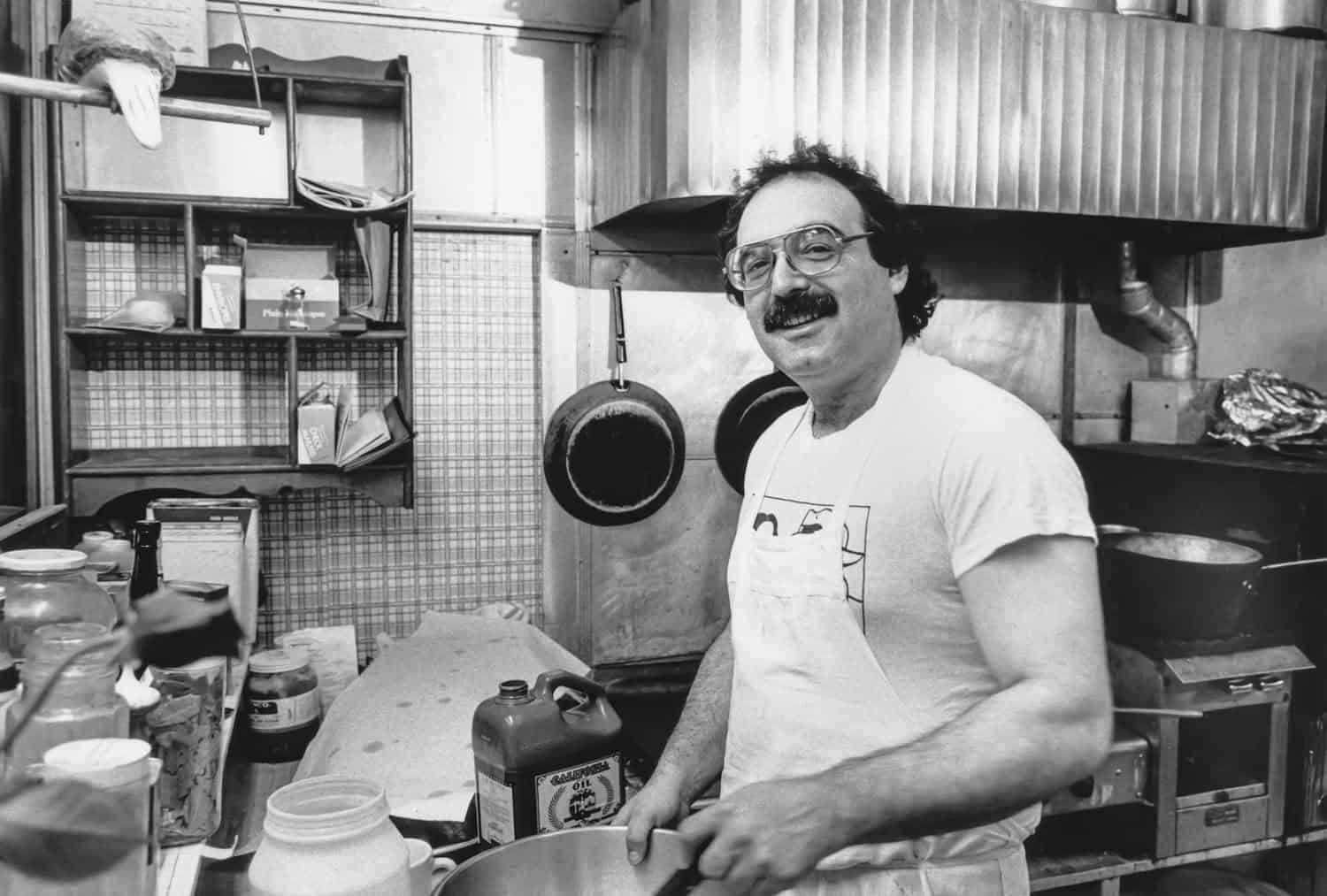 Black-and-white photograph of a middle aged man with dark balding hair and a dark mustache. He has on a white apron and appears to be in a kitchen. He smiles at the camera.