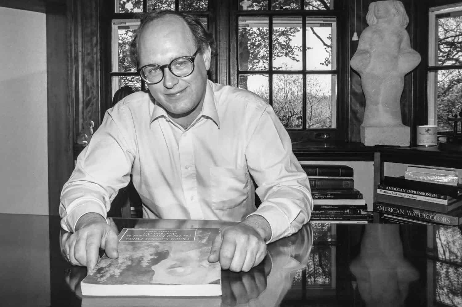 Black-and-white photograph of a middle aged man sitting infront of a window. He has balding light brown hair and glasses. He is smirking at the camera and resting his hands on a book. He has on a white buttoned down shirt.
