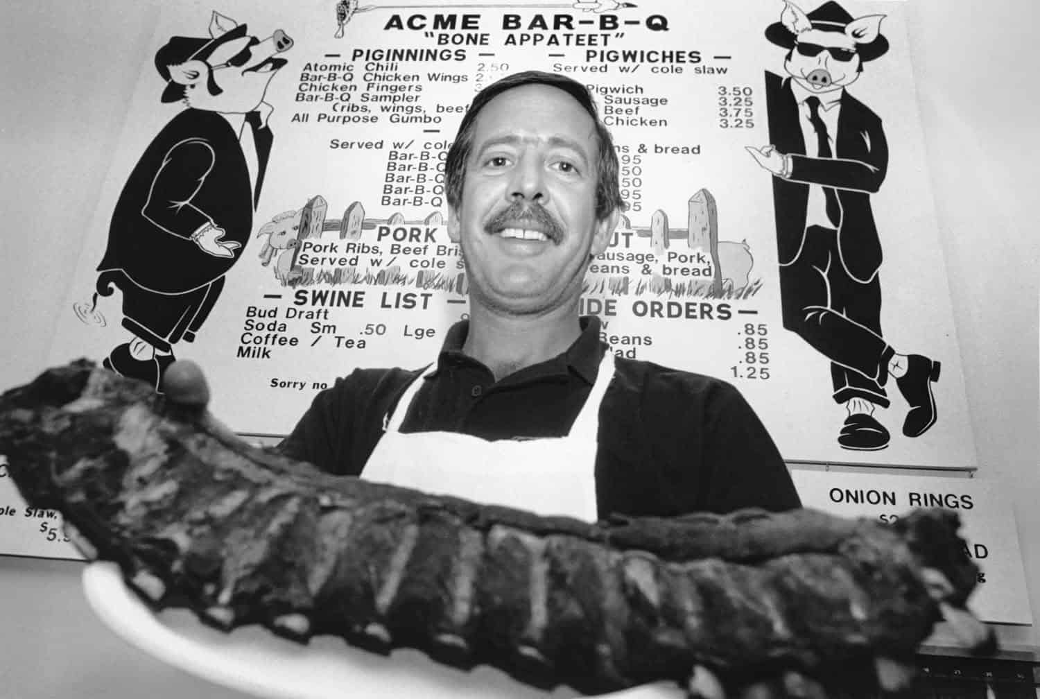 Black-and-white photograph of a middle aged man holding meat toward the camera. He is smiling. He has short dark hair and a dark mustache. He is wearing a black collared shirt with a white apron on it. A menu for "ACME BAR-B-Q" featuring two anthropomorphic pigs in suits and sunglasses.