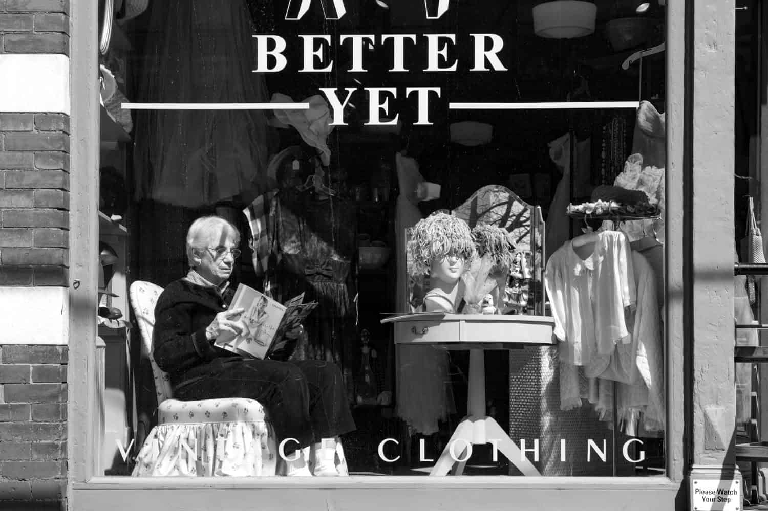 Black-and-white photograph of a large window wall of a storefront. On the window it reads: "K & J BETTER YET." A matured woman is sitting infront of the window reading a magazine.