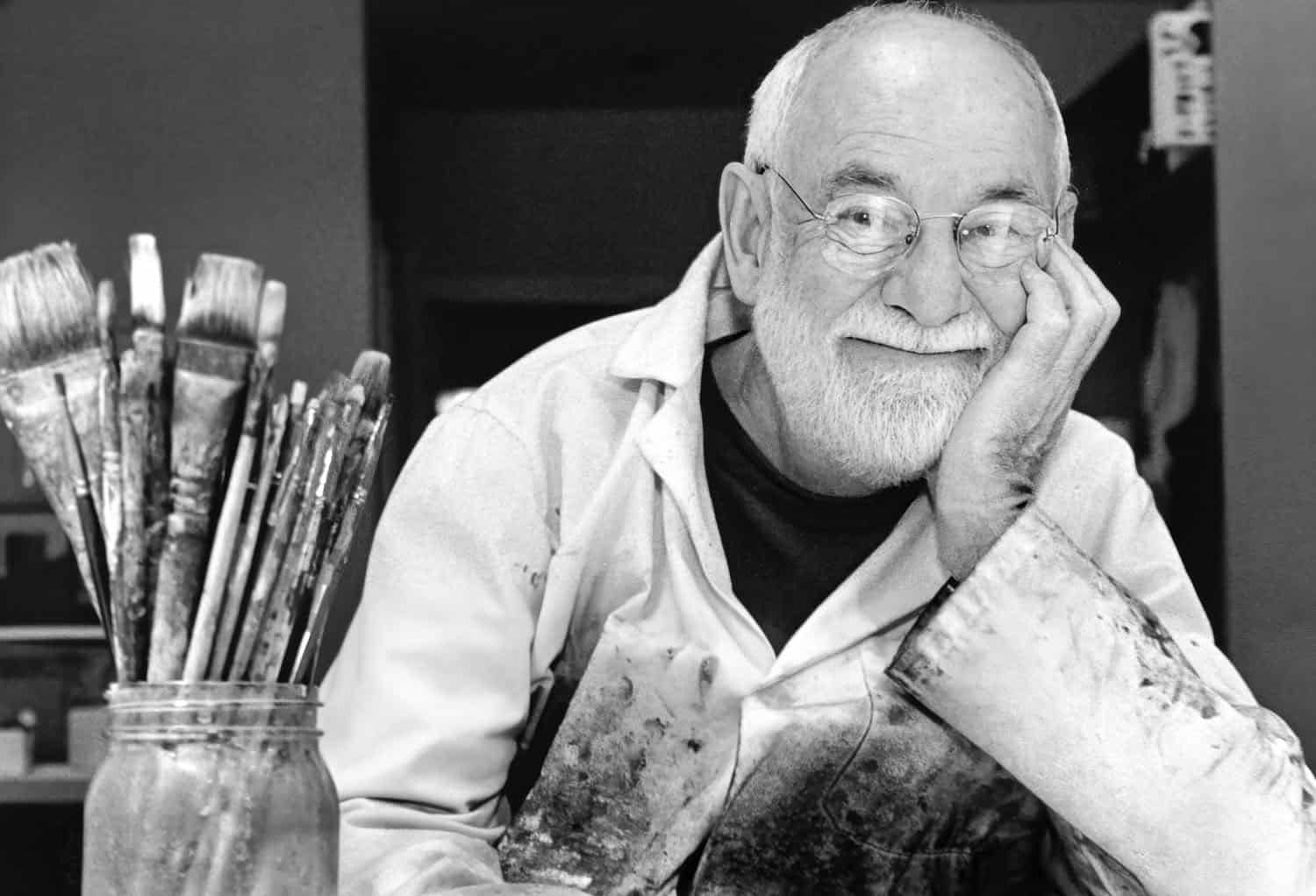Black-and-white photograph of a matured Eric Carle. He has white balding hair and a white beard. He is wearing oval glasses and resting his hand on his hand. Next to him is a cup of paint brushes.
