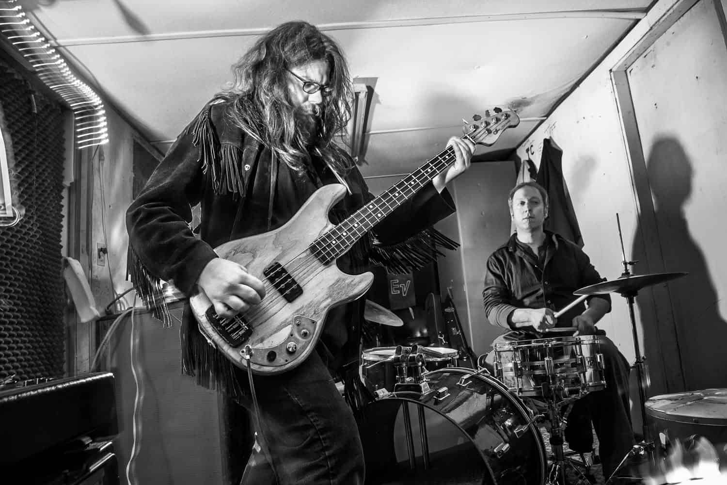 Black-and-white photograph of a bearded man with long hair playing the guitar while another man plays drums in the background.