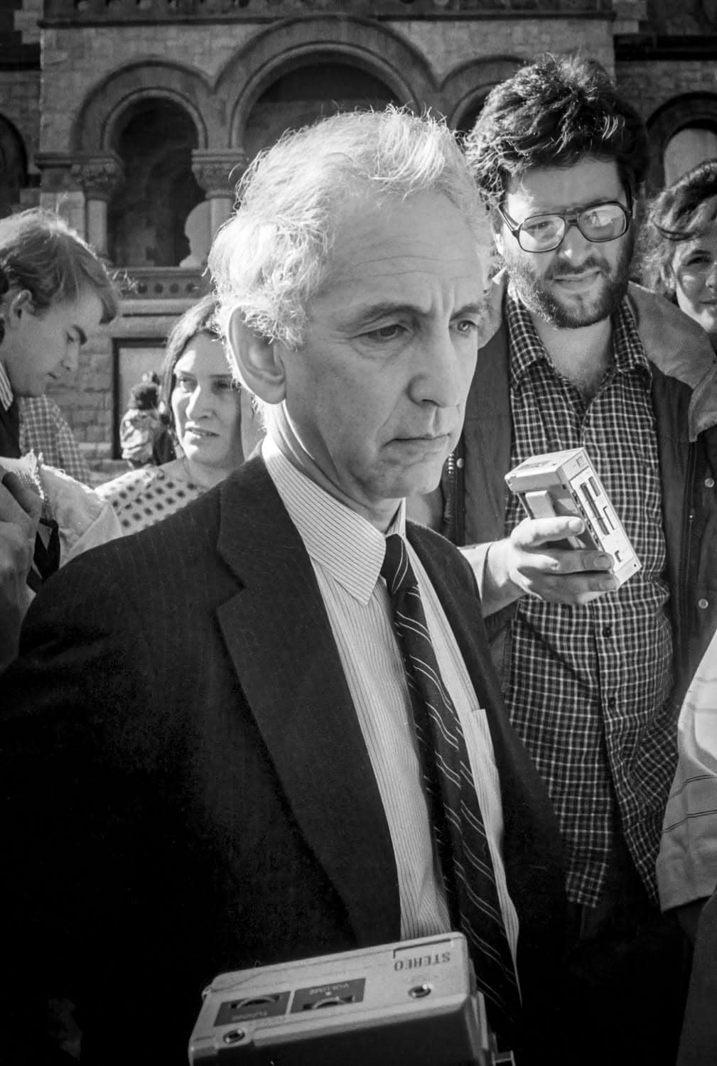 Photograph of a middle-aged man standing infront of the courthouse on Main Street (Northampton). A crowd is gathered behind him. He has a saddened or worried expression as he looks off into the distance. He is wearing a black and white suit and tie and has white hair.