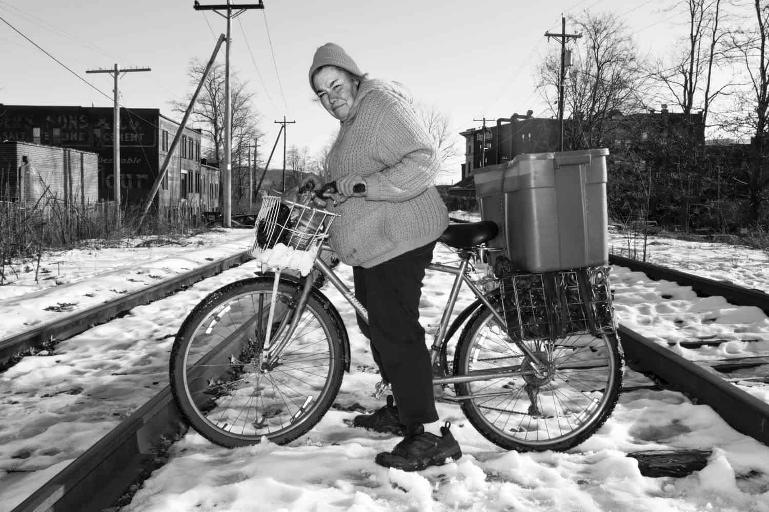 Black-and-white photograph of a middle-aged woman on a bicycle. On the back of the bicycle is a cart holding a box. There is snow on the ground. 