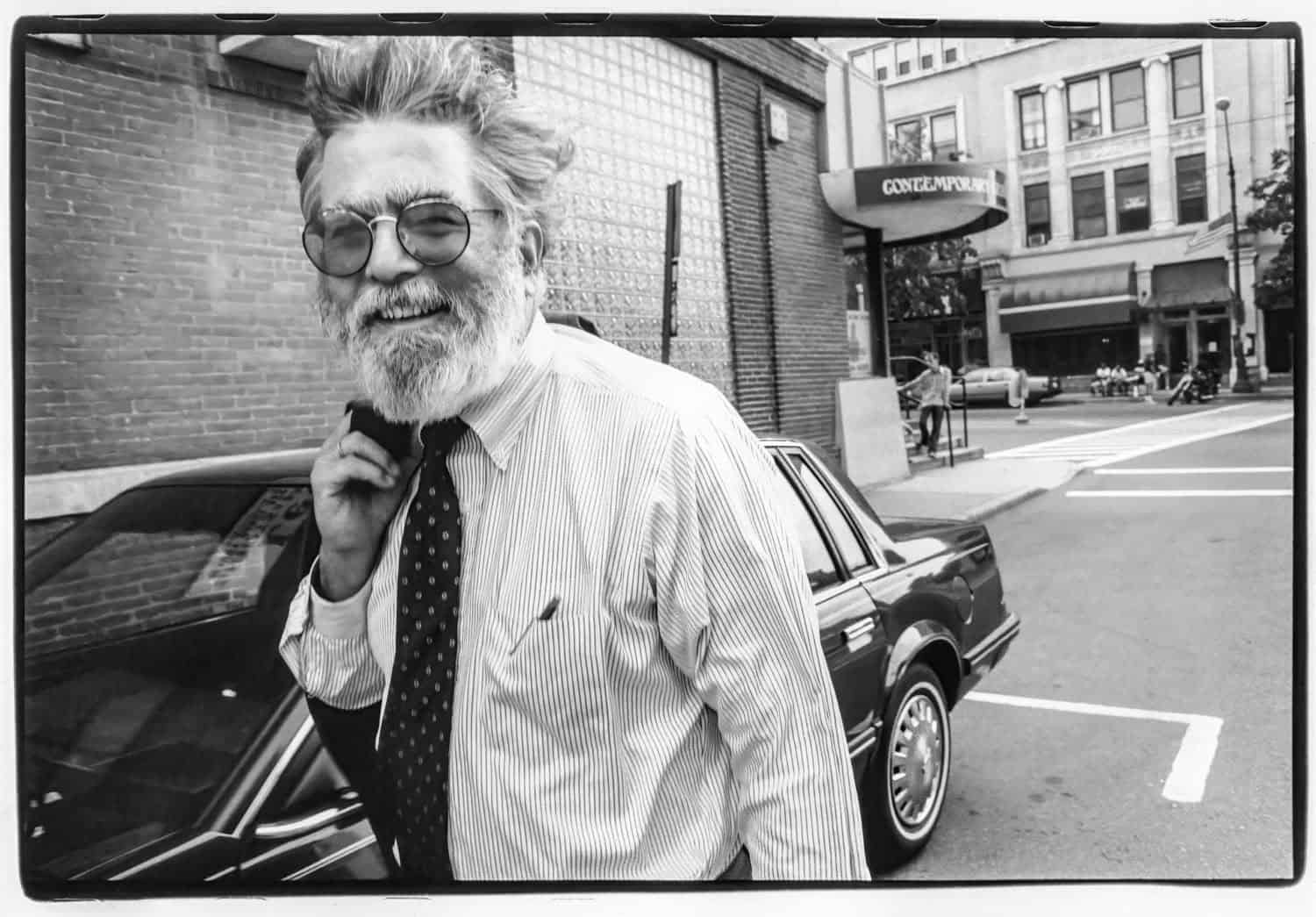 Black-and-white photograph of a man walking down a side road off of Main Street, Northampton. He is wearing a striped suit and a poka-dotted tie. He is holding something over his shoulder. He has graying white hair and a beard and is wearing glasses. He smiles at the camera.