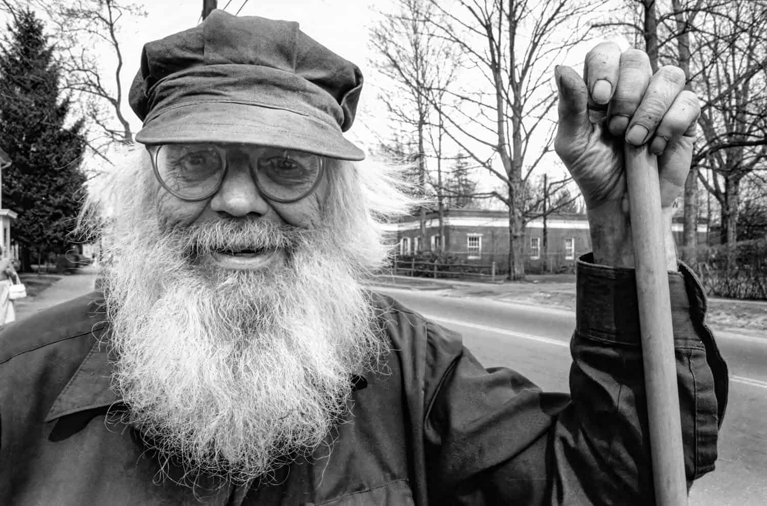 Photograph of a matured man on Bridge Street. He is wearing a hat and has a long white beard and glasses. He is holding onto a gardening tool.