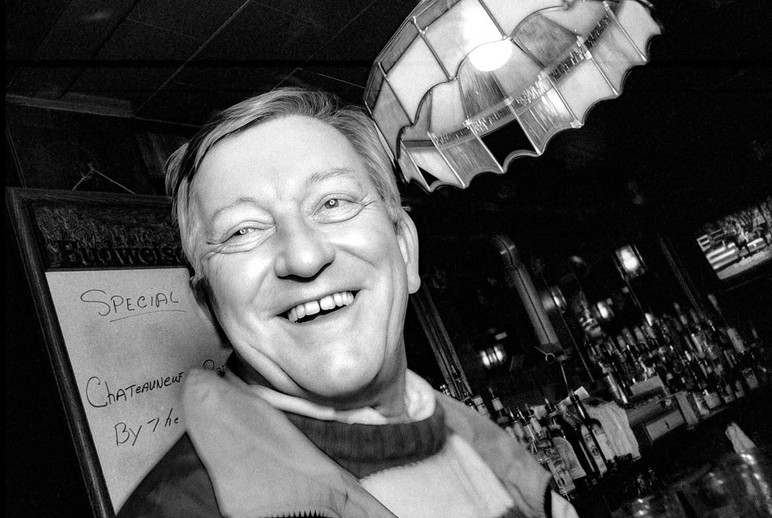 Black-and-white photograph of a matured man smiling at the camera. He appears to be sitting at a bar or pub. He has white hair.