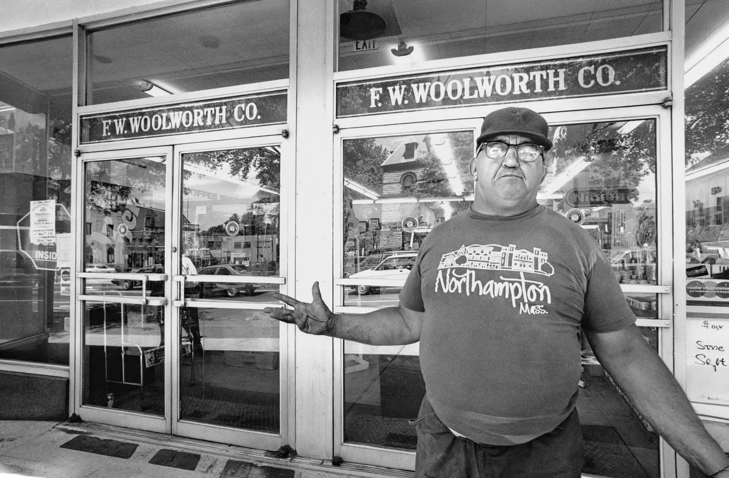 Black-and-white photograph of a man standing infront of a storefront "F.W. WOODWORTH CO." He has his arms out in an expressive way and is wearing a Northampton t-shirt and baseball cap.