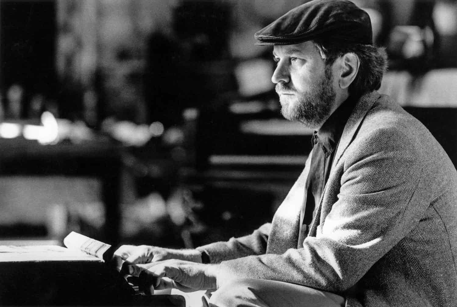 Photograph of a middle aged man playing a small keyboard. He has on a black cap and a gray suit jacket. He is bearded. He looks off to the side. The background is blurred.
