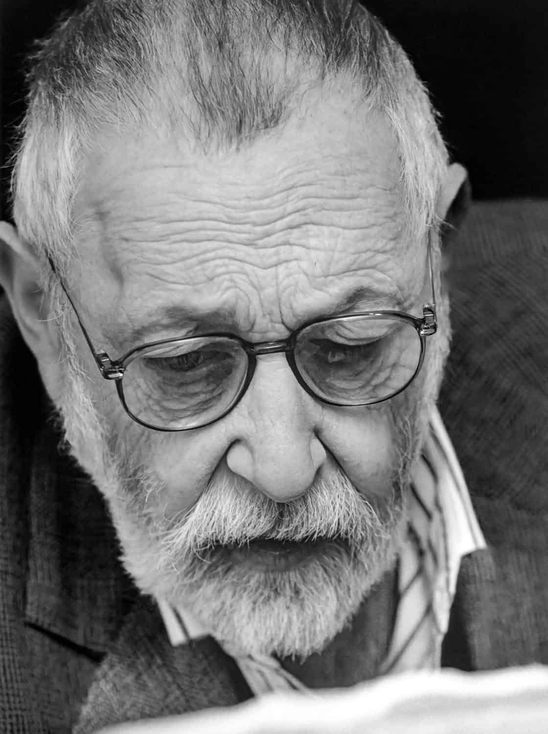 Black-and-white photograph of a matured man with white hair and beard and glasses. He looks down at a book.