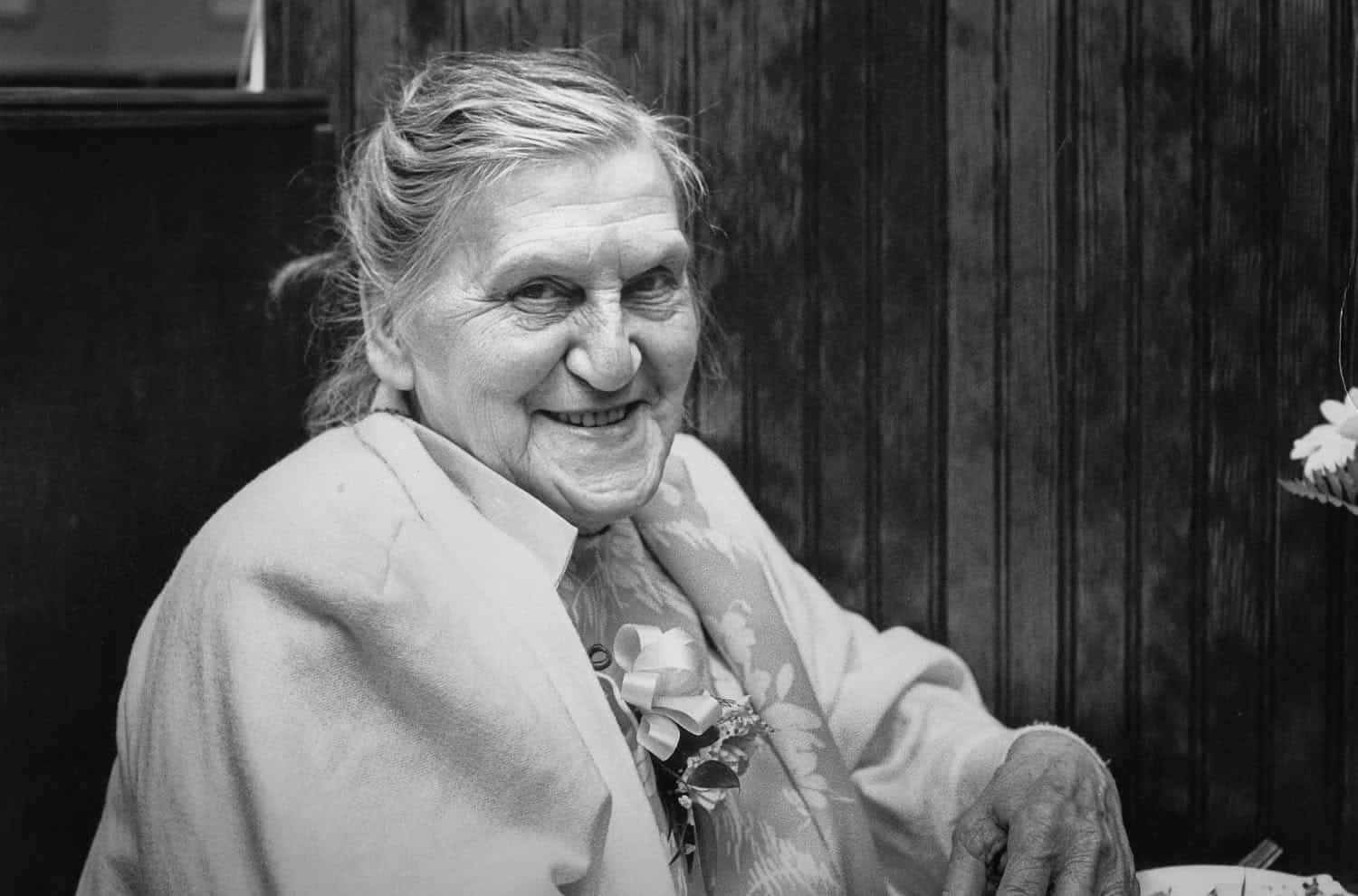 Black-and-white photograph of a matured woman smiling at the camera. A wooden wall is behind her. She has white tied back hair and is sitting down with her face turned toward the camera.