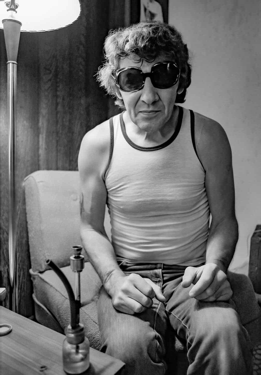 Black-and-white photograph of a middle aged man with crooked sunglasses on. He has short curly brown hair and is wearing a white tank top and jeans. He is sitting on a  cushined chair with a lamp next to him.