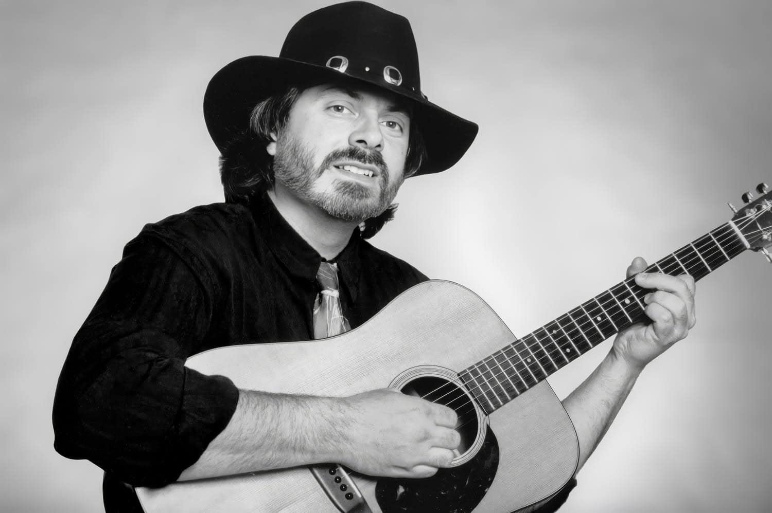 Photograph of a man in a black suit with a lighter tie holding a guitar as if he is beginning to play. He has a neutral, almost smiling expression on his face and has a beard. On his head is black cowboy hat and the background is light and solid.