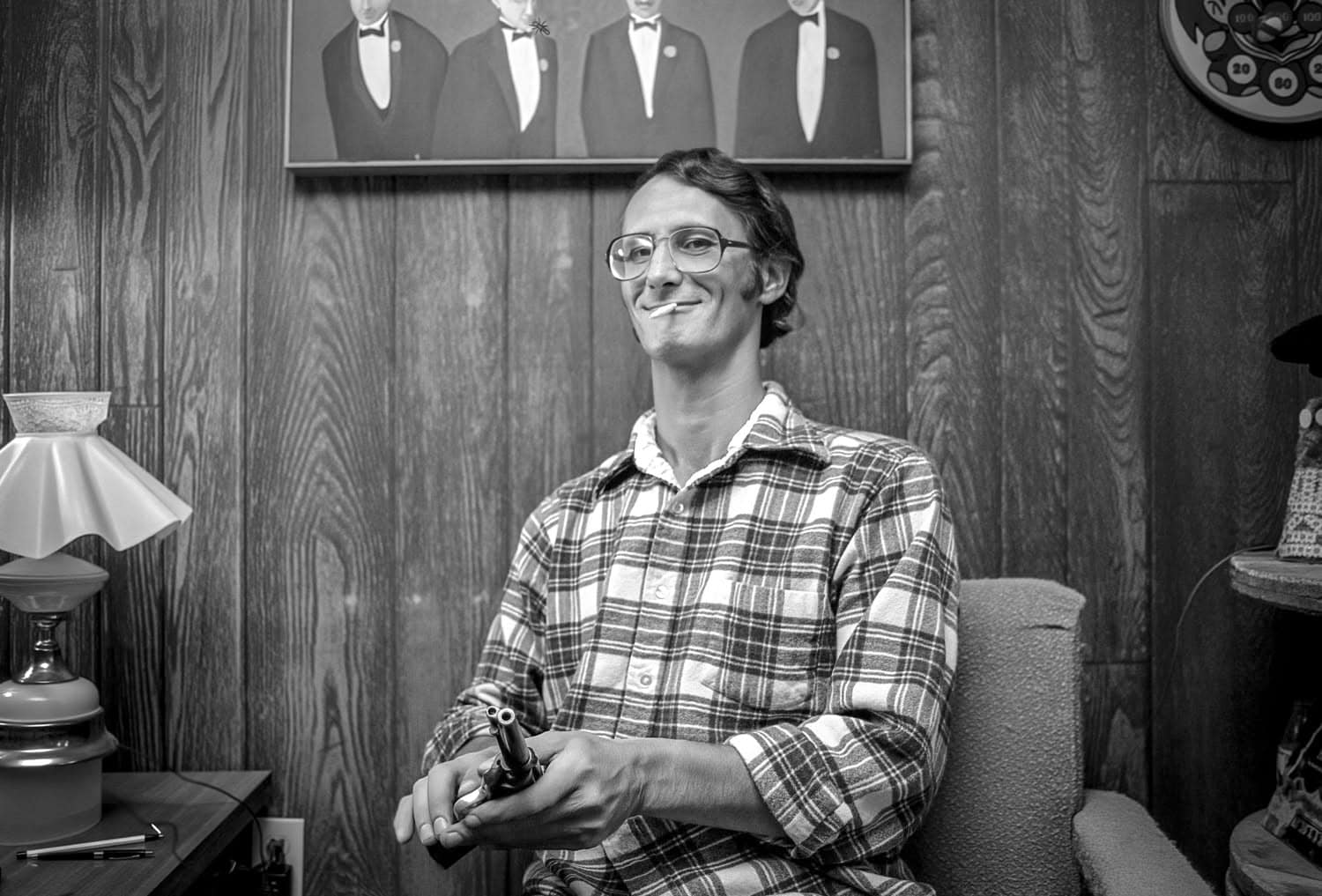 Black-and-white photograph of a young man sitting in a chair. He has short hair and glasses and is wearing a plaid button down shirt. He is smiling with a cigarette in his mouth.