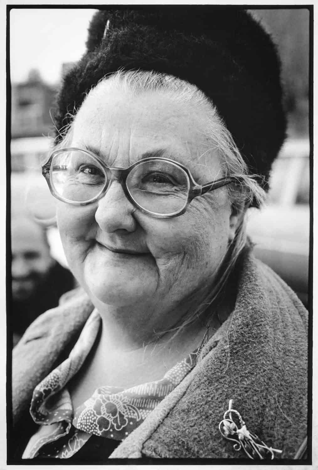 Black-and-white portrait photograph of a matured woman smiling at the camera. She wears round glasses and a fuzzy black hat. Her graying hair peaks through the hat. The background is blurred.
