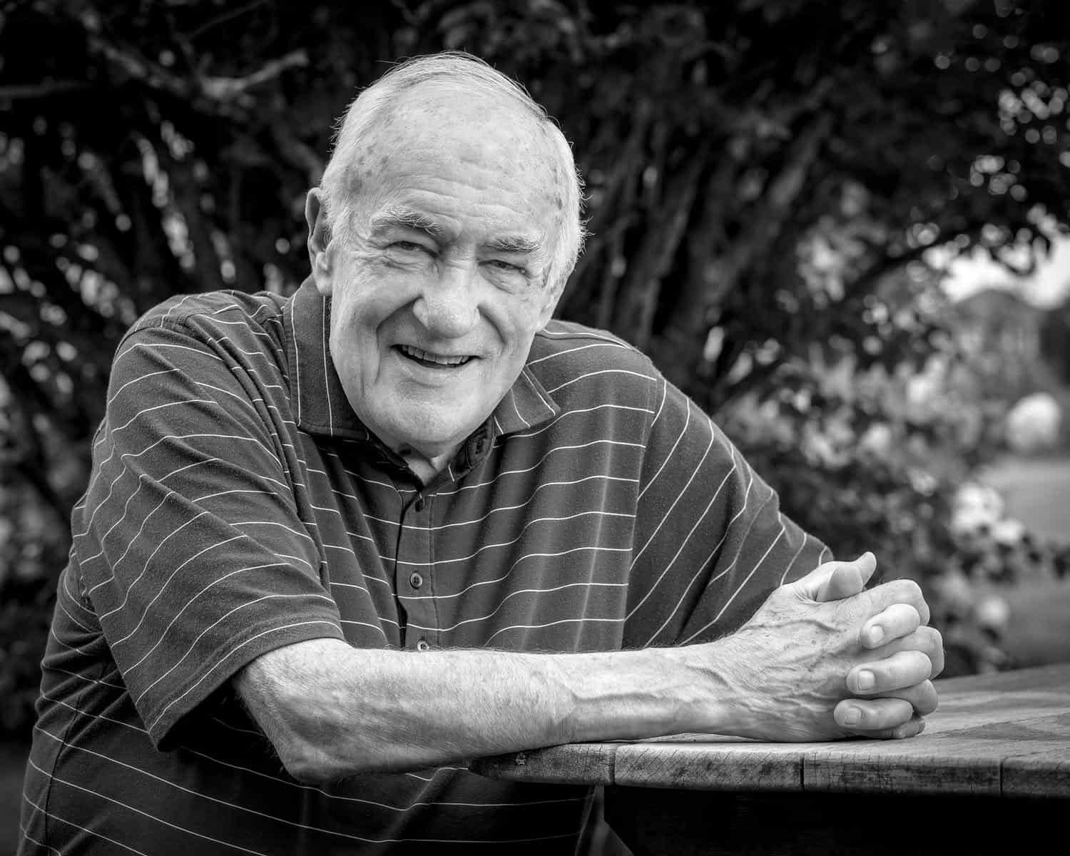 Photograph of a matured man smiling at the camera. He is in a striped button down shirt and is holding his hands together resting them on a table. In the background is a tree.