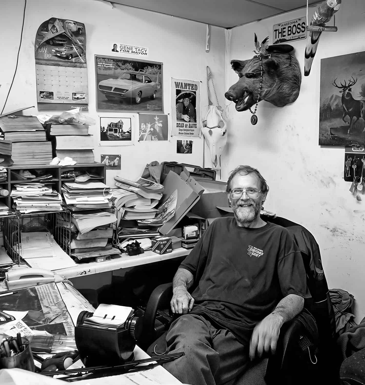 Black-and-white photograph of a middle aged man with dark but graying hair and beard. He is sitting at a cluttered table or desk.