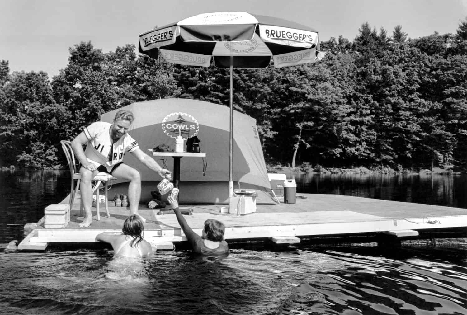 Black-and-white photograph of a young man sitting on a platform on the water in a chair under an umbrella handing drinks to people swimming in the water.