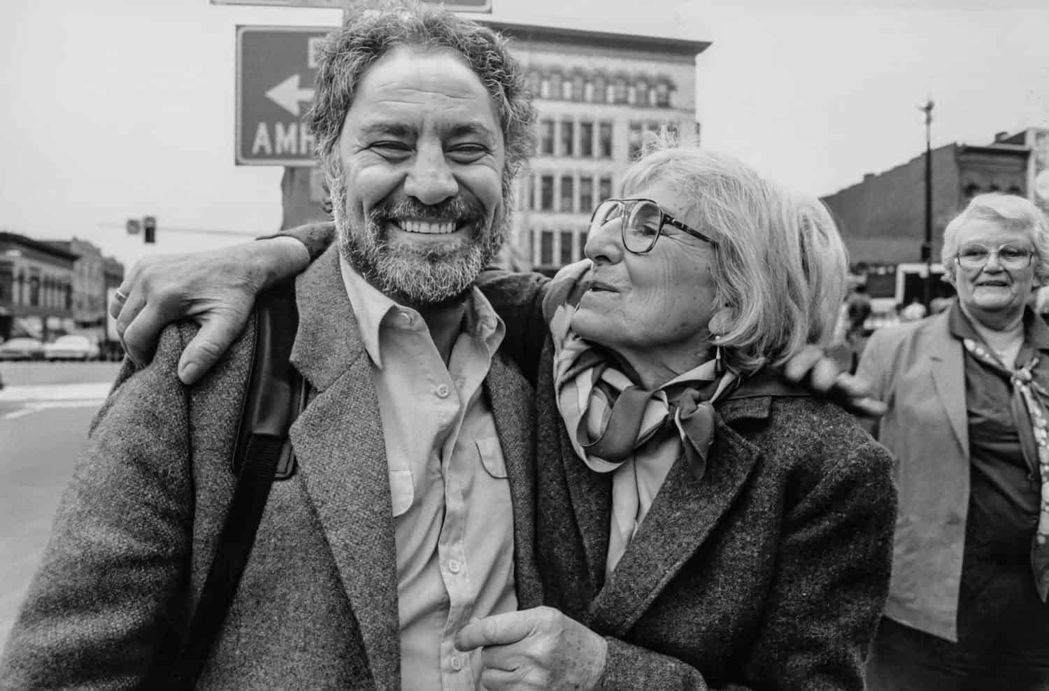 Photograph of a middle aged man and matured woman on Main Street. The man is smiling at the camera and the woman is embracing him and looking at him.