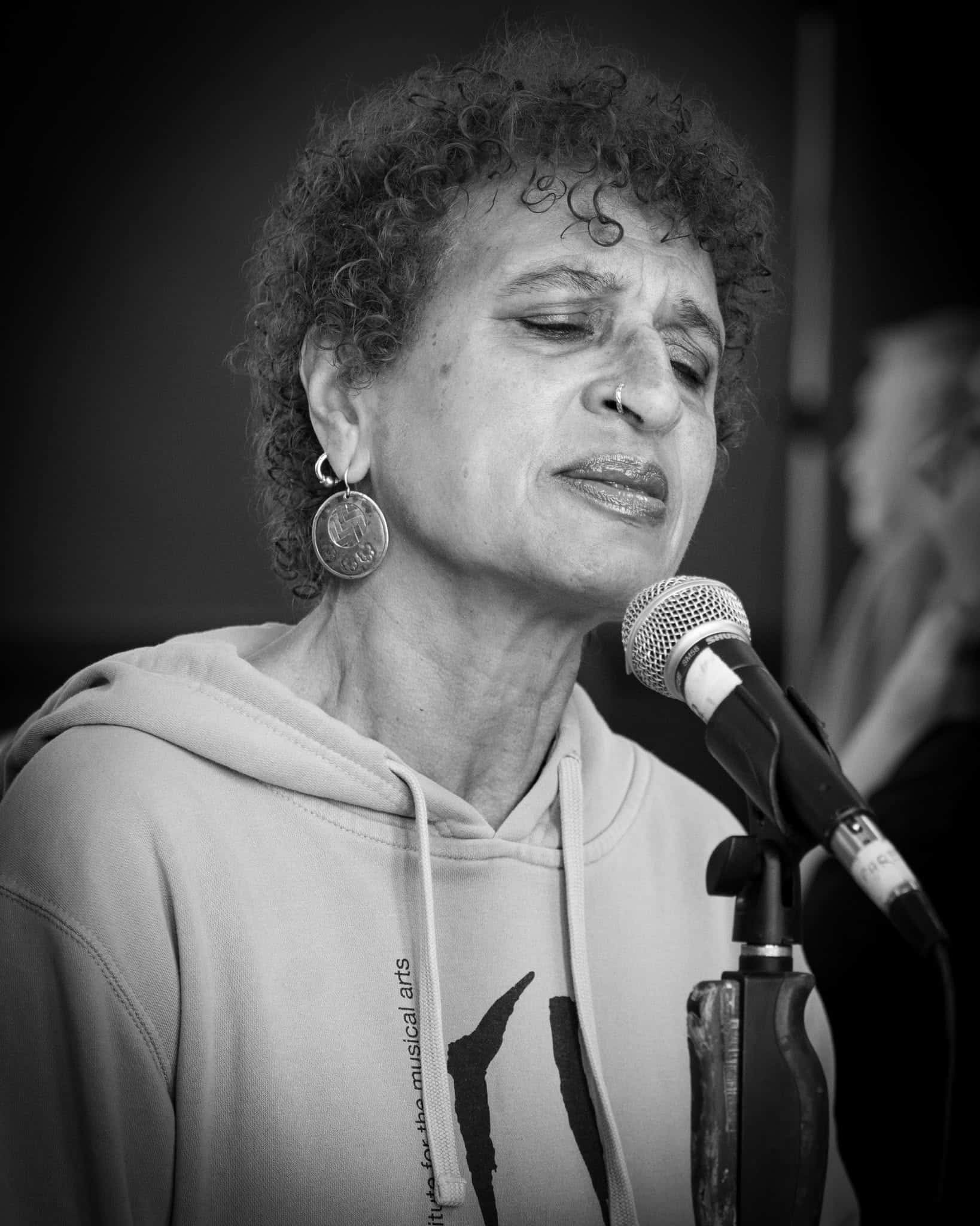 Photograph of a matured woman singing into a microphone with her eyes closed. She is wearing a hooded sweatshirt and has on round dangle earrings. Her hair is short and curly, and she has a nose piercing with hoop jewelry. 