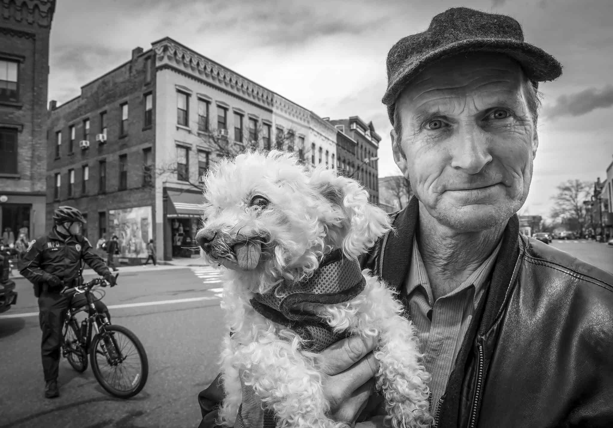 Photograph of a matured man on Main Street in Northampton. He is holding a small white dog that is sticking out its tongue. The man is wearing a buttoned down shirt and a leather jacket. He has on a baseball cap and is smirking at the camera.