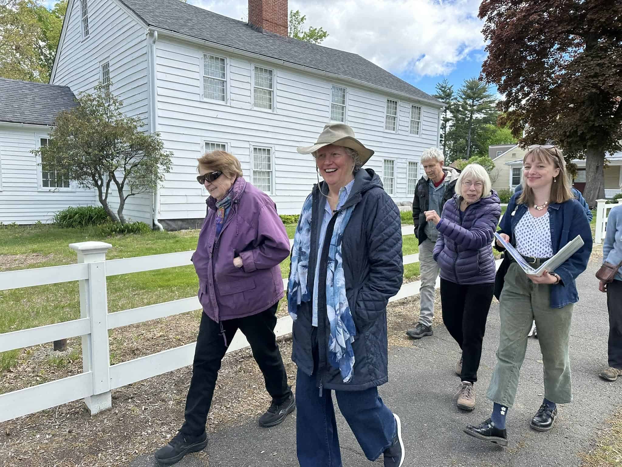 Photograph of a group of people walking past an 18th century house with white siding and a red brick chimney with a white picket fence out front. The crowd is being led by a young woman with shoulder length blonde hair, wearing a jean jacket, green pants, black boots, and a white top. She is holding a white binder and smiling at the camera.