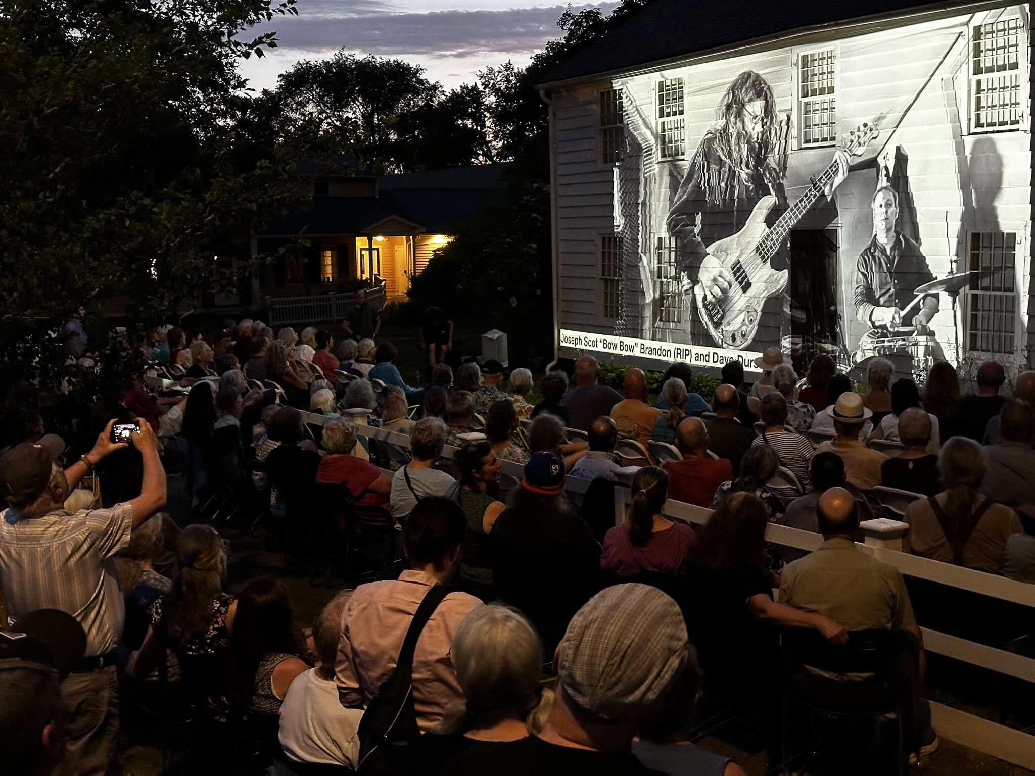 People gathering in front yard of Parsons House at night. Photographs from "Faces of Northampton" projected on the house.