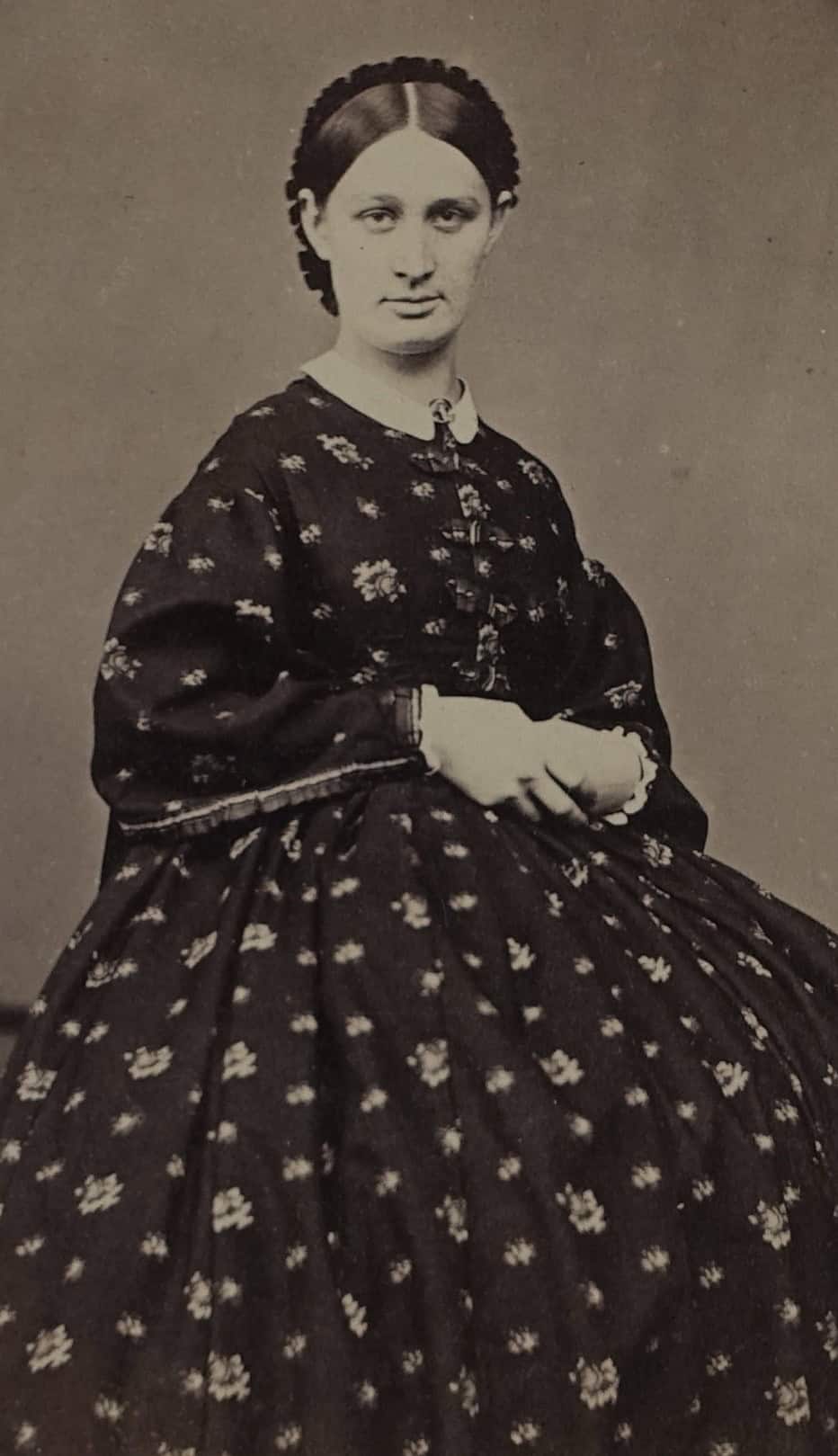 Black-and-white photograph of a young woman in her late 20s or 30s sitting on a chair and posing with her hands placed together on her lap. She is wearing a dark colored-floral dress with a white babydoll collar.