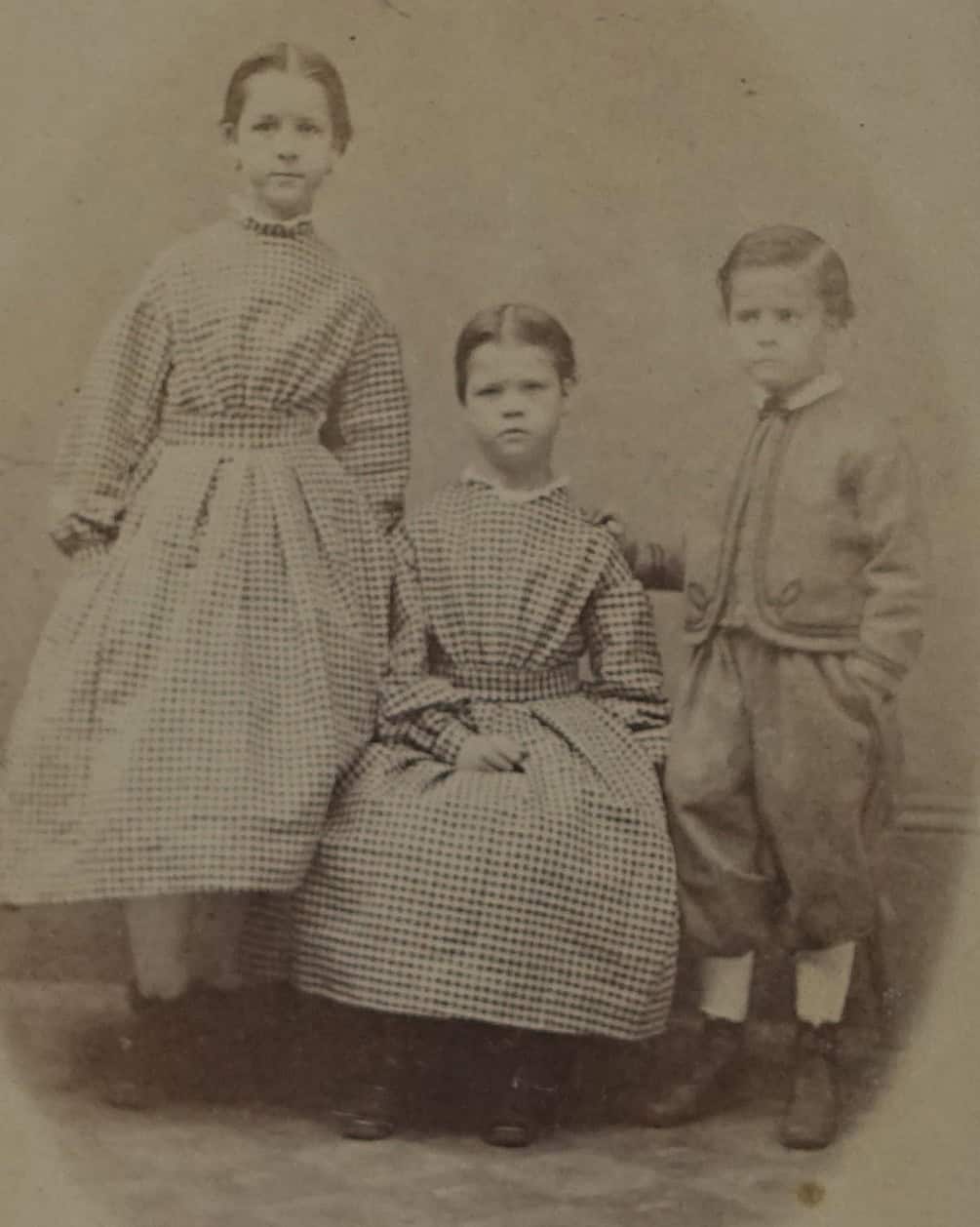 Black-and-white photographic portrait of three young children, On the right, is a little girl of elementary school age standing with a plaid dress. In the middle is a younger girl, still around elementary school age, sitting with an identical plaid dress on. The left most child is a little boy, an older toddler, standing with dress paints and a jacket.