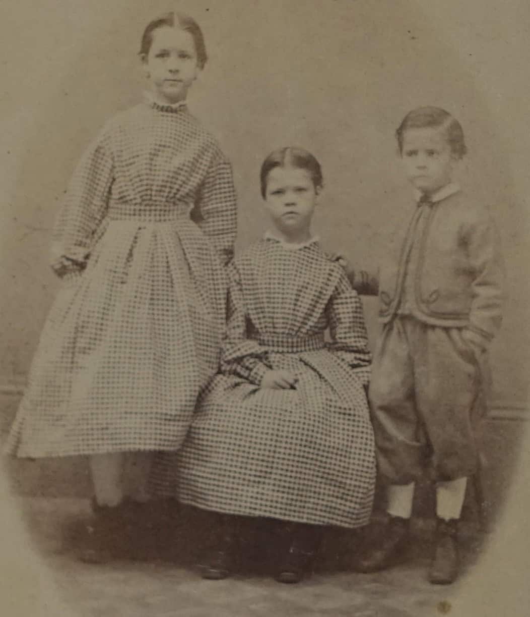 The image shows three children in an old, yellowed photograph. It captures a young girl standing on the left side of the frame. She has her hair pulled back smoothly and wears a long-sleeved, high-collared dress with a dark checkered or plaid pattern. The dress features a fitted bodice and a full, pleated skirt that reaches below her knees, revealing light-colored stockings and dark boots. To the right of her, another girl sits down wearing a nearly identical checkered dress, while a young boy stands on the far right wearing a short jacket, trousers, and light-colored socks with boots.