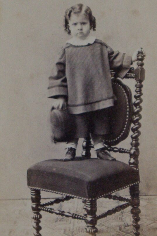This black-and-white photograph depicts a young child, standing upright on the upholstered seat of an ornate wooden chair. The child has dark, shoulder-length hair styled in soft curls or ringlets and is looking directly at the camera with a neutral, serious expression. They are dressed in period clothing consisting of a loose, long-sleeved, knee-length tunic or smock featuring a prominent white collar, worn over short trousers or bloomers. On their feet are dark, buttoned ankle boots with light-colored socks. The child's left hand rests on the decorative backrest of the chair for stability, while their right hand holds a dark, rounded hat down by their side.