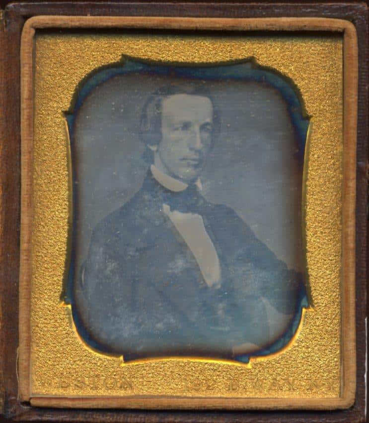A bust-length portrait of a clean-shaven man with dark, slicked-back hair. He is facing slightly to his right, wearing a dark suit jacket, a white collared shirt, and a dark bowtie. The image is encased within a textured, gold-colored wood frame.