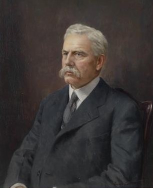 A painting depicting the upper body of a mature man with white hair and a prominent, full white mustache. He is turned slightly to the left, wearing a dark gray or charcoal-colored suit jacket over a white collared shirt and a muted gray tie. The background is a solid, dark brown color, and he appears to be sitting in a dark wooden chair.