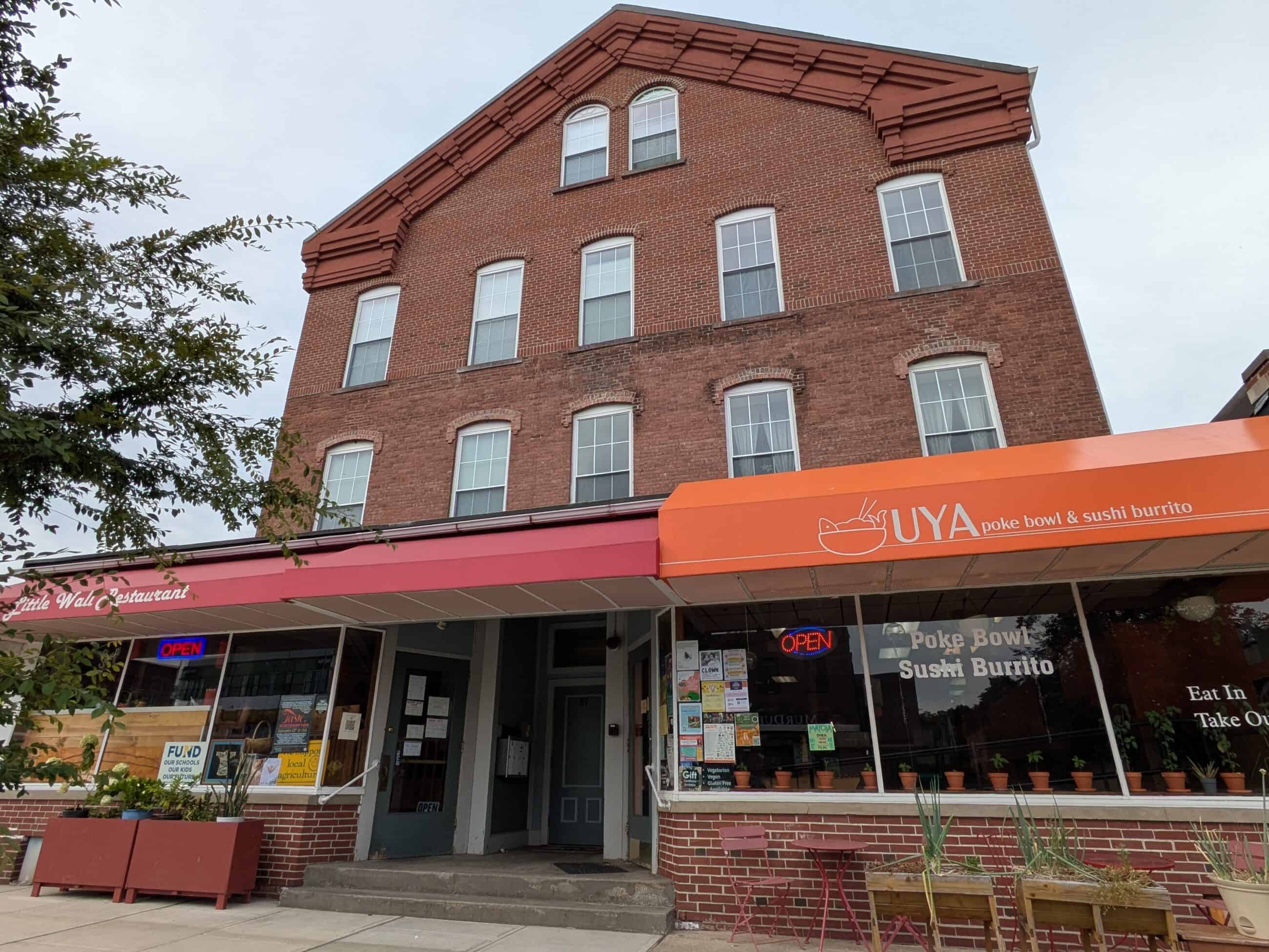 Image of red brick building with small more modern front addition with marquee covers reading "Little Wali Restaurant" and "UYA poke bowl & sushi burrito." Many posters posted on the front windows.