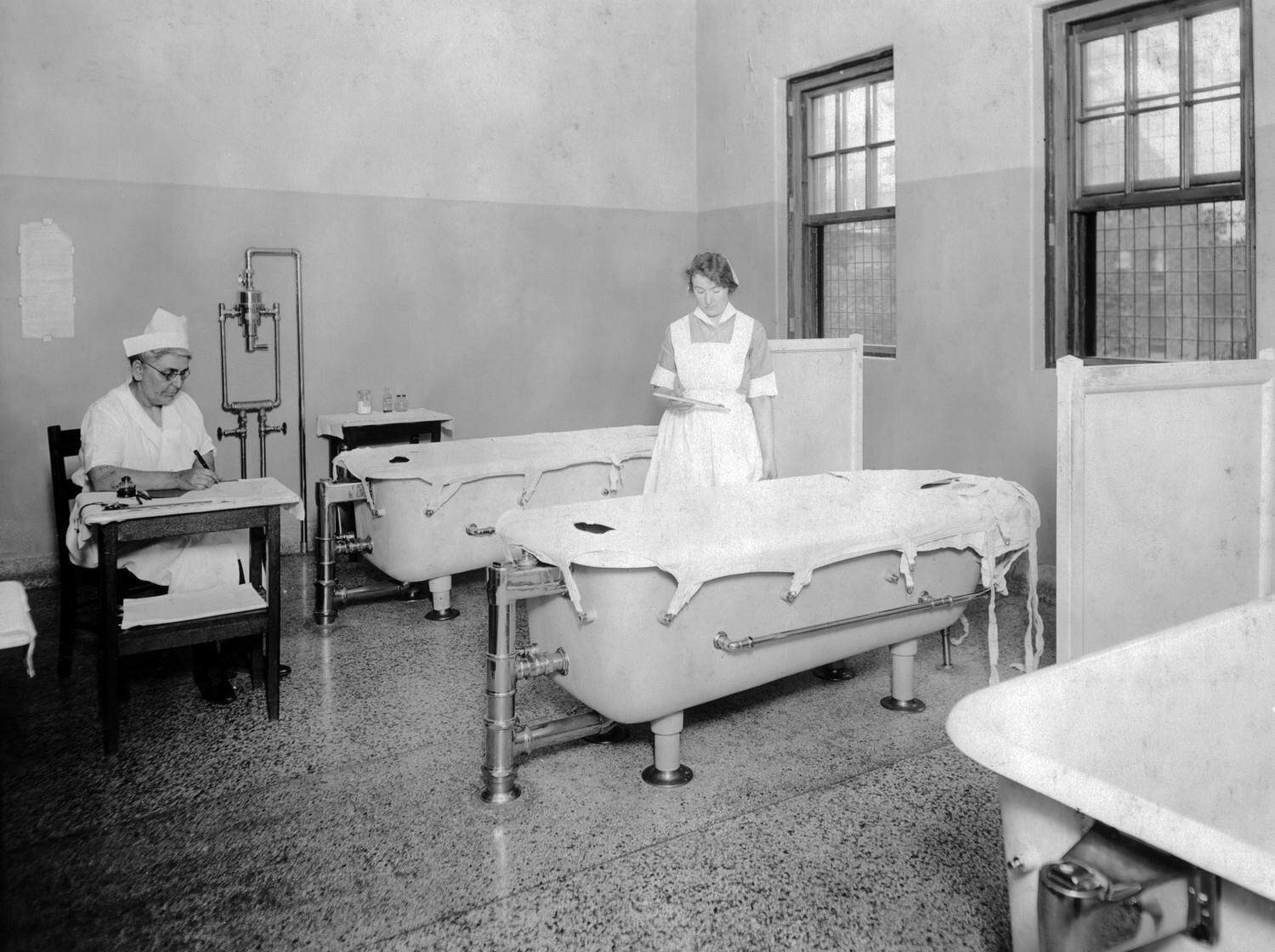 Black-and-white photograph of two nurses among several large therapeutic bathtubs with canvas covers, tiled partitions, tall windows, and polished floors, documenting the hospital's hydrotherapy facilities.