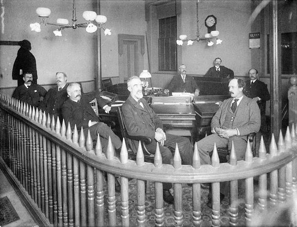 A black and white photograph depicting a group of nine men in formal attire gathered in a council chamber. In the foreground, a prominent, curved wooden balustrade with distinctive, pointed picket-like finials sweeps across the lower portion of the frame. Behind this barrier, three men sit closely together on the left, while two men occupy large chairs near the center. The man in the center-left position has gray hair, a full white beard, and looks toward the camera. To his right, another man in a light gray suit sits facing forward. In the background, four additional men are positioned behind a long wooden desk or bench arrangement. The room features high ceilings, a hanging multi-bulb light fixture, a wall clock, and a paneled door.